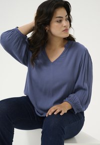 Blusa leggera di colore blu chiaro, con una scollatura a V e maniche a palloncino arricciate. Abbinata a jeans in denim scuro. Focalizzata su comfort e vestibilità.