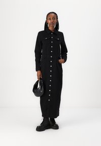 Robe chemise en denim noir avec des manches longues, un col, des boutons à l'avant et des poches latérales ; associée à des bottes à lacets noires et un sac rond.