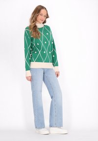 Maglione verde con un motivo geometrico bianco e accenti di texture, abbinato a jeans a zampa d'elefante blu chiaro e sneakers bianche.