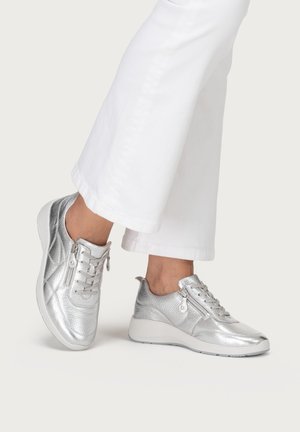 Füße, die silberne Metallic-Sneaker mit seitlichen Reißverschlüssen und Schnürsenkeln tragen, kombiniert mit weißen, weiten Hosen vor einem einfarbigen Hintergrund.
