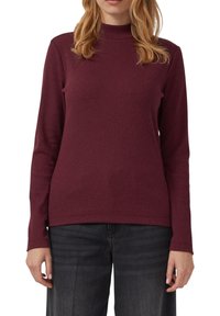Vrouw in een bordeauxrode langmouw turtleneck trui en zwarte jeans, staand tegen een witte achtergrond.
