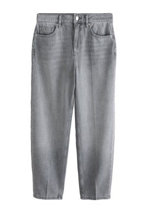 Ung kvinde går fremad iført en hvid oversized t-shirt med blonderkant, grå jeans og sølv sneakers mod en ensfarvet lys baggrund.