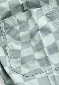 Das karierte Gewebe zeigt verschiedene Grüntöne und Grautöne. Es beinhaltet eine aufgesetzte Tasche mit sichtbaren Nähten und einer glatten Textur.