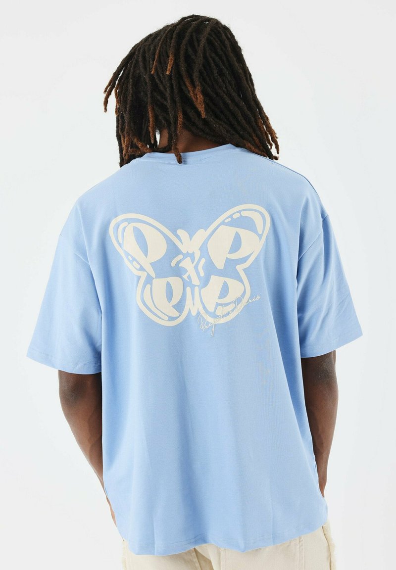 Camiseta de algodón azul claro con un gran gráfico de mariposa color marfil en la parte posterior, con letras estilizadas y un detalle de firma.