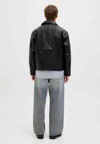 Schwarze Lederjacke mit strukturiertem Design, ausgestattet mit einem spitzen Kragen und einer geraden Silhouette. Getragen über einem grau gestreiften Hemd und lockeren Jeans.