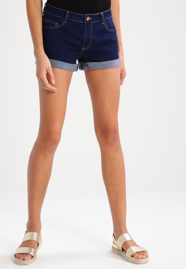 Vrouw draagt donkerblauwe opgerolde spijkershorts, zwart topje en gouden sandaaltjes met riempjes, staat met licht gespreide benen op een witte achtergrond.