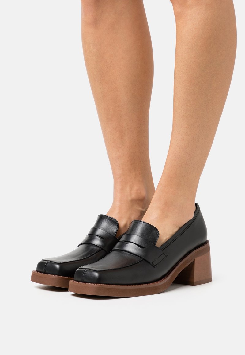 Jambes inférieures d'une femme portant des mocassins à talons carrés en cuir noir avec semelles marron sur fond blanc.