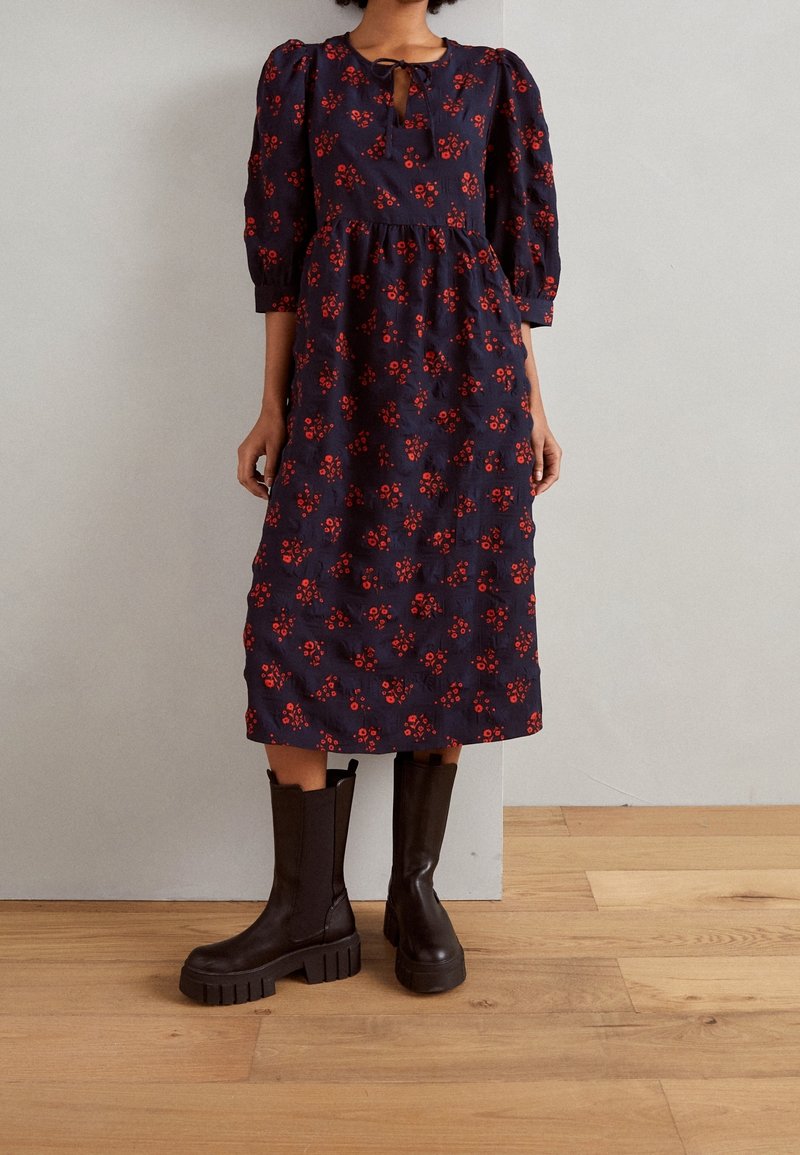 Femme portant une robe midi bleu marine à motif floral rouge, manches bouffantes et bottes noires jusqu’aux genoux, debout à l’intérieur sur un sol en bois.