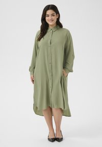Abito verde oliva a maniche lunghe con bottoni e orlo asimmetrico, tasche laterali e maniche arrotolate. Indossato con tacchi a punta neri.