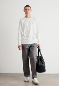 Langermet hvit skjorte med en liten logo, grå bukser og hvite sneakers; holder en svart bag, minimalistisk design, innendørs setting.
