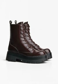 Cizme scurte din piele maro cu șiret, eyelets întărite și o talpă groasă de culoare neagră. Include bucle de prindere pe tijă.