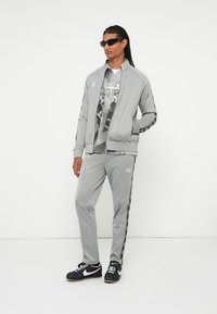 Chaqueta de chándal gris con pantalones a juego, presenta detalles y logotipo en negro. El modelo lleva una camiseta gráfica y gafas de sol. Zapatos deportivos completan el look.