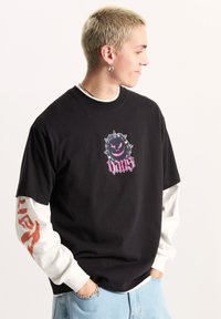 Camiseta negra de manga corta con un diseño gráfico en rosa y negro que presenta una cara sonriente y picos, superpuesta sobre una camiseta blanca de manga larga.