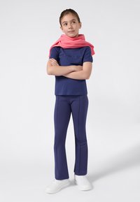 Jong meisje staat met gekruiste armen, gekleed in een marineblauwe Champion-outfit, witte sneakers en een roze sweatshirt over haar schouders.