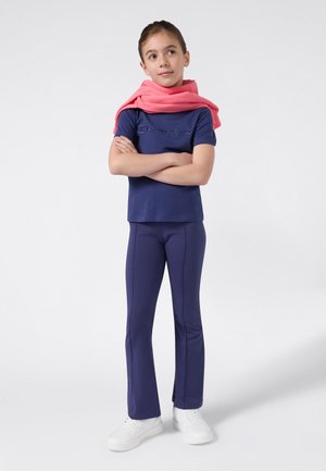Jong meisje staat met gekruiste armen, gekleed in een marineblauwe Champion-outfit, witte sneakers en een roze sweatshirt over haar schouders.