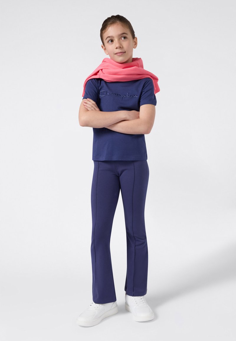Jong meisje staat met gekruiste armen, gekleed in een marineblauwe Champion-outfit, witte sneakers en een roze sweatshirt over haar schouders.