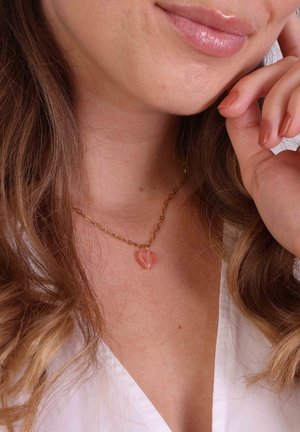 Gouden ketting met een hartvormige rozekleurige kristallen hanger. Heeft een gestructureerd schakelontwerp en ligt op de huid met los bruin haar.