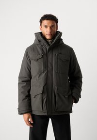 Giacca isolante verde scuro con cappuccio, multiple tasche frontali, maniche lunghe e chiusura con zip. Realizzata in tessuto resistente e strutturato.