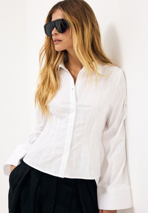 Kobieta z długimi blond włosami, nosząca czarne, duże okulary przeciwsłoneczne, białą teksturowaną koszulę na guziki i czarne spodnie, stoi na tle białej ściany.