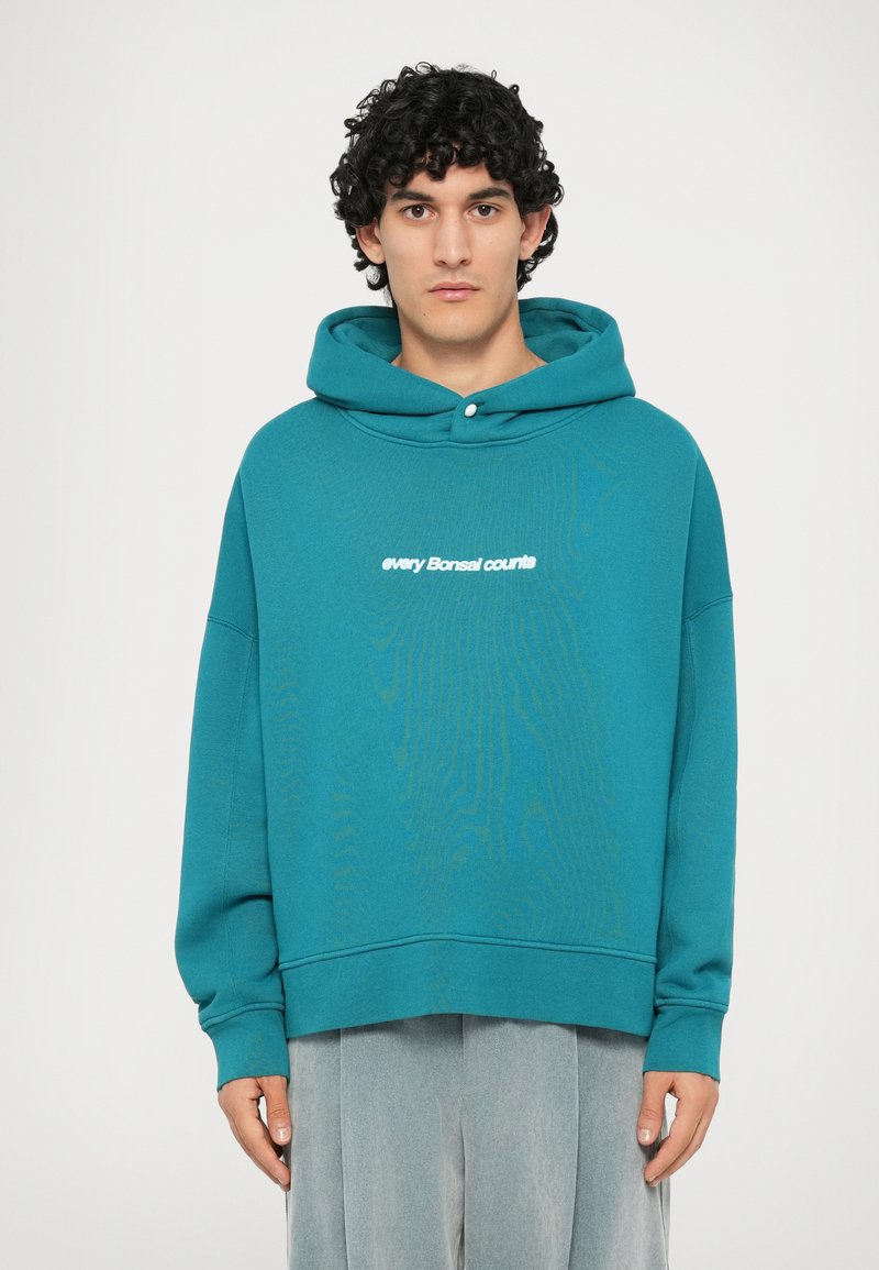 Türkise Kapuzenpullover aus weichem Material, mit lässiger Passform und weißem Text "Jeder Bonsai zählt" auf der Vorderseite.