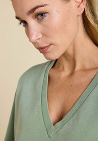 T-shirt à col en V en tissu vert doux et léger. Présente des manches courtes et une texture subtile. La vue rapprochée met en évidence l'encolure et le teint de la peau.