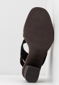Semelle d'une sandale noire avec une bande de roulement texturée et un talon en caoutchouc antidérapant appuyé contre une surface blanche à angles.