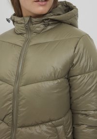 Veste rembourrée vert olive avec capuche, dotée d'une fermeture éclair, d'une texture matelassée et d'accents en métal. Légère et isolante.