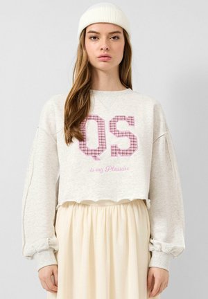 Junge Frau mit weißer Mütze, beigem kurz geschnittenem Sweatshirt mit "QS" und plissiertem cremefarbenem Rock, stehend vor einem schlichten hellgrauen Hintergrund.