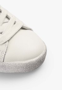 Gros plan sur l'avant d'une basket blanche avec une semelle en caoutchouc texturée gris clair et des lacets blancs sur un fond blanc uni.