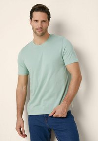 Tricou verde mentă, cu guler rotund, dintr-un material de bumbac moale, cu mâneci scurte ajustate, purtat cu blugi bleu închis. Design simplu, casual.