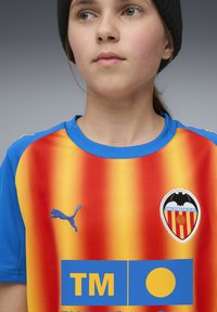 Ung person som bär en Valencia CF fotbollströja med orange och röda ränder, blå ärmar, en Puma-logotyp och klubbens emblem synligt.