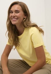 T-shirt jaune à manches courtes en tissu doux avec un col rond ; offre une coupe décontractée et une texture lisse. Le modèle est assis dans un environnement neutre.