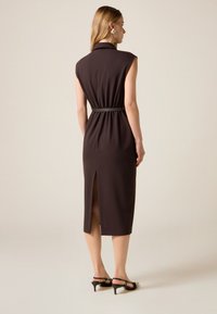 Brun ærmeløs kjole med bælte i taljen, knælængde med slid bagpå og glat tekstur. Sorte stropper hæle fuldender looket.