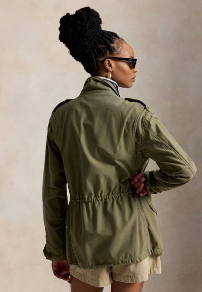 Vrouw met gevlochten haar in een knot, zonnebril, een olijfgroene jas en beige korte broek, staand tegen een neutrale achtergrond in zijaanzicht.
