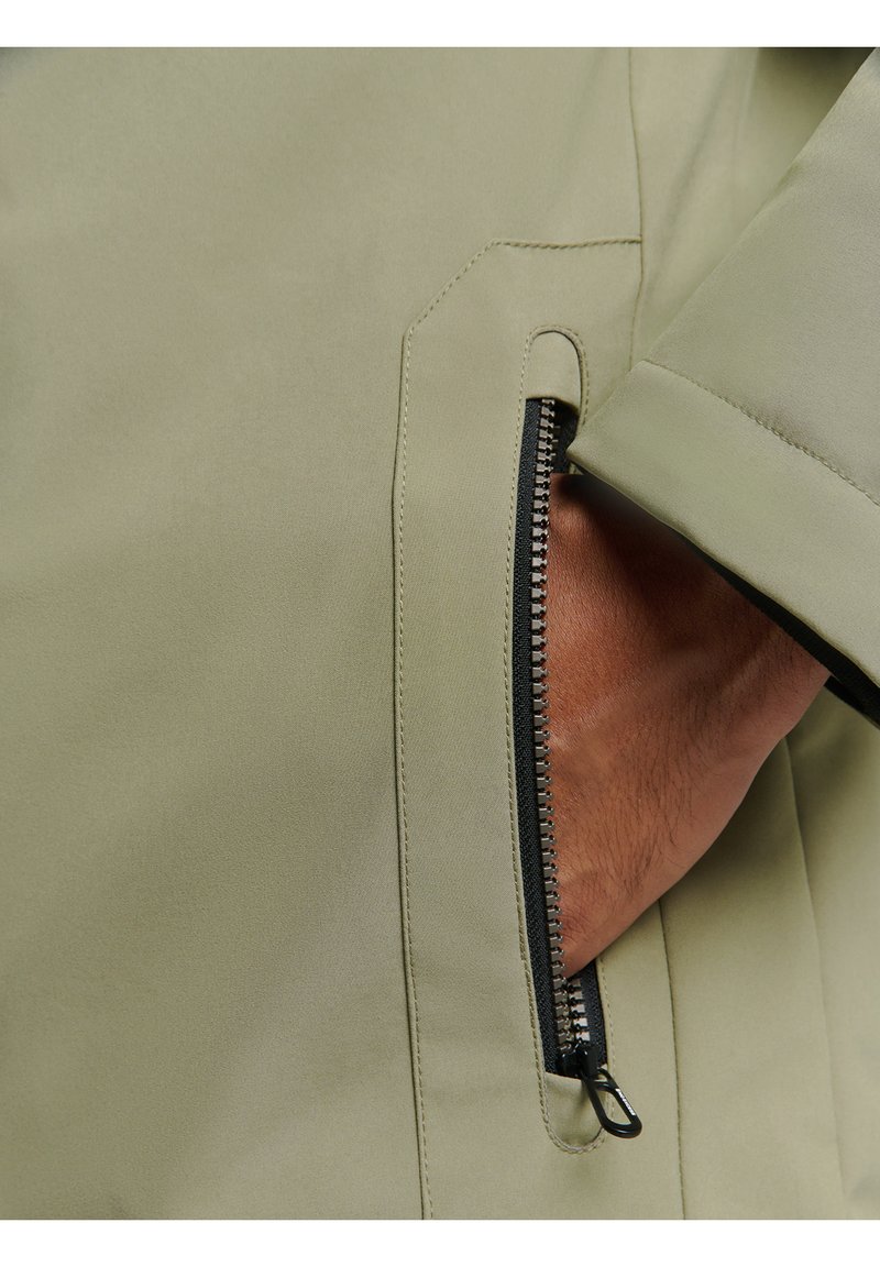 Veste vert clair avec une poche zippée sur le côté ; finition lisse et mate ; fermeture éclair et accents noirs sur les bords ; vue en gros plan de la texture et des détails.