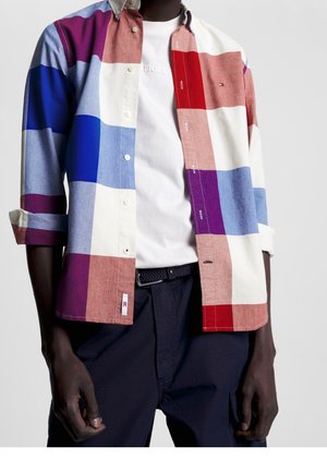 Homme portant une chemise à carreaux multicolores boutonnée par-dessus un t-shirt blanc et un pantalon foncé, avec les manches retroussées.