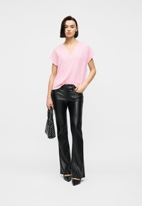 Γυναίκα που φοράει ροζ υφασμένο τοπ με κοντά μανίκια, μαύρο δερμάτινο παντελόνι, μαύρες μυτερές γόβες, κρατώντας μια μαύρη τσάντα με σχέδιο.