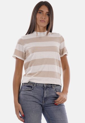Donna con lunghi capelli castani che indossa una maglietta beige e bianca a righe a maniche corte e jeans blu, in piedi con una mano in tasca.