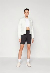 Hvit jakke med glidelås og strukturert finish, kombinert med svarte skreddersydde shorts og lyse sneakers. Modellen står foran en ensfarget bakgrunn.