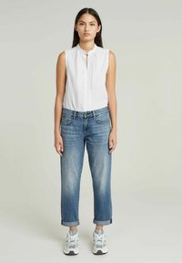 Camisa sin mangas blanca con rayas verticales, cuello con botones, combinada con jeans azul claro de puños enrollados y zapatillas deportivas grises.