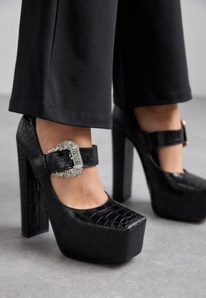 Zwarte krokodillen-embossed hoge hakken met een chunky plateau en een decoratieve zilveren gesp op een enkelbandje. Glad oppervlak, strakke uitstraling.