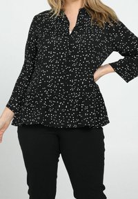 Blusa peplum negra con lunares blancos, cierre de botones en la parte delantera, mangas largas y tela texturizada. Combinada con pantalones negros.
