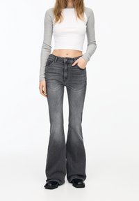 Pantalones vaqueros grises acampanados con una textura de denim suave, cintura alta, bolsillos frontales y ligeros tonos desgastados, combinados con una camiseta de manga larga blanca y gris.