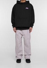 Musta huppari, jossa on violetti logo, hevosenkenkätasku ja rento istuvuus, yhdistettynä vaaleanpuhuvaan leveälahkeisiin housuihin ja mustiin tennareihin.