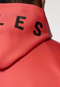Sweat à capuche rouge en tissu texturé, avec de grandes lettres noires au dos et une finition lisse.