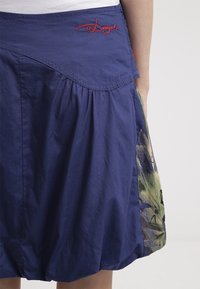 Jupe bleu marine avec tissu froncé, logo rouge brodé "Desigual" sur la ceinture et panneau en tissu fleuri sur le côté.