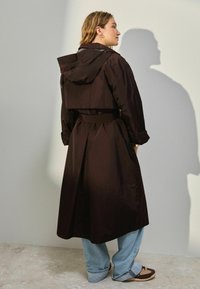 Mørkebrun regnfrakke med hætte, indsnøret talje og mellemlang design. Har en bælte-lukning og store sidelommer.