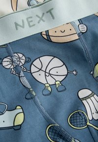 Tissu bleu avec des illustrations ludiques sur le thème du sport, comprenant un ballon de basket souriant, une raquette de tennis, un volant de badminton et une chaussure, avec une ceinture grise portant l'inscription "NEXT".