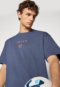 Mørkeblå bomull t-skjorte med rund hals, løstsittende. Har et oransje "INTER" logo og en Nike swoosh-emblem over det. Spilleren holder en hvit fotball.