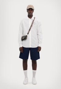 Camicia bianca a maniche lunghe con bottoni, pantaloni corti blu navy, calze chiare e sneakers bianche. Cappellino beige con cinturino e una piccola borsa a tracolla nera.